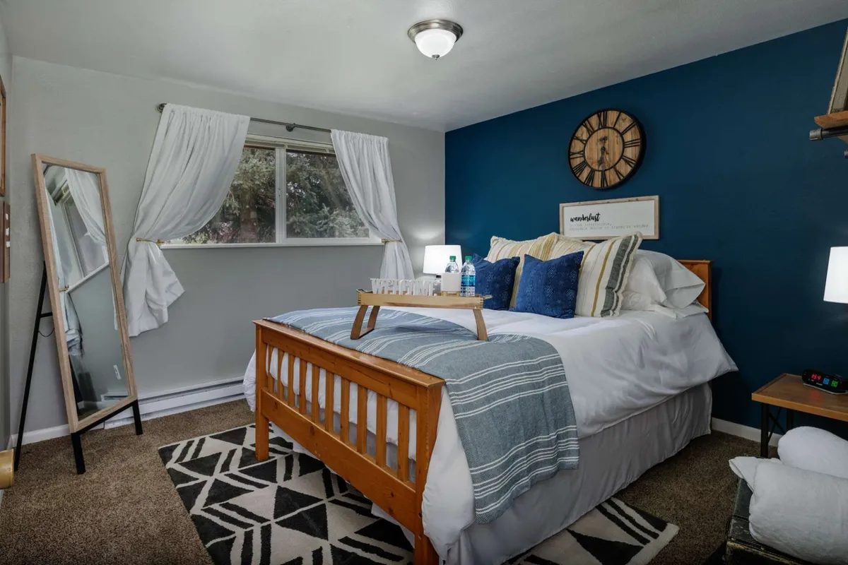 This image showcases a serene and inviting bedroom with a queen-sized bed as the focal point, accented by a stylish blue wall, decorative clock, and a mirror. The bed appears plush and welcoming, exuding a sense of comfort and tranquility ideal for restful nights or romantic getaways. The space is bright with natural light, and tasteful décor elements create an elegant atmosphere.
