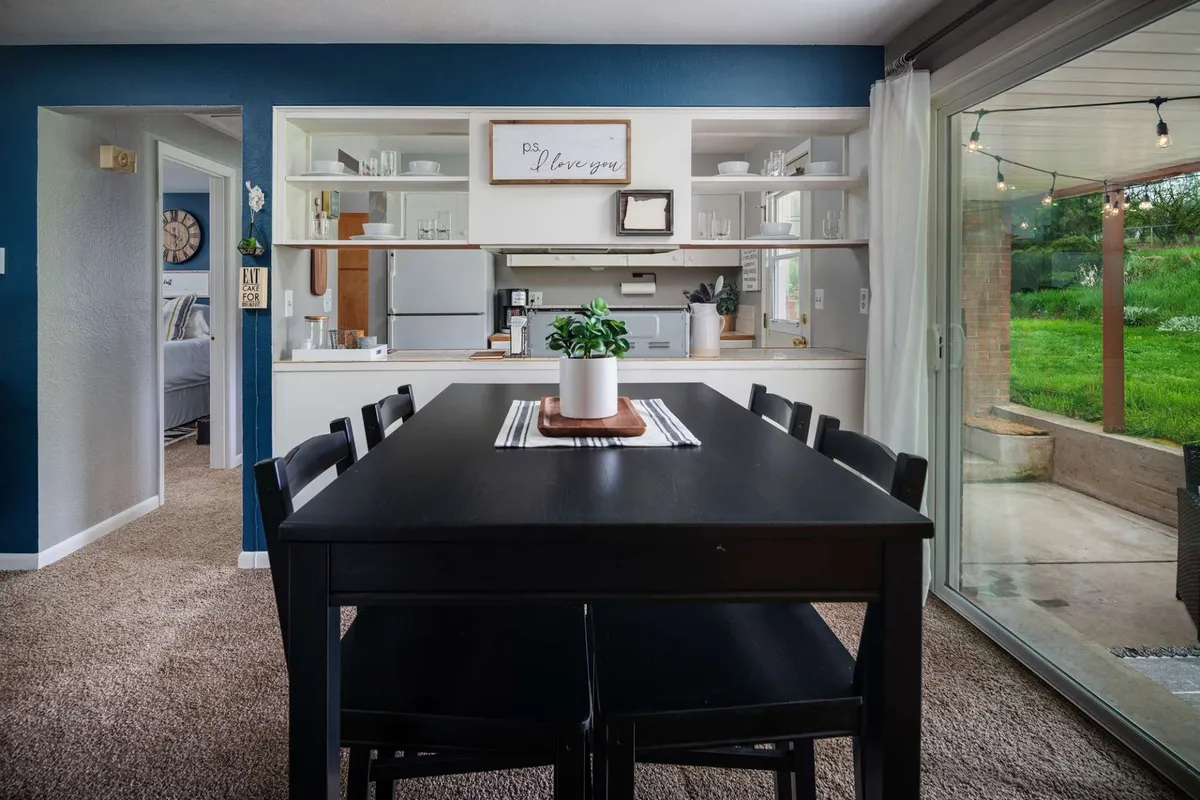 This image shows an inviting dining table set within an open-plan space, with kitchen shelves visible in the background and a prominent sliding glass door that brings in natural light. The setup suggests a fluid connection between dining, cooking, and enjoying the view outside.