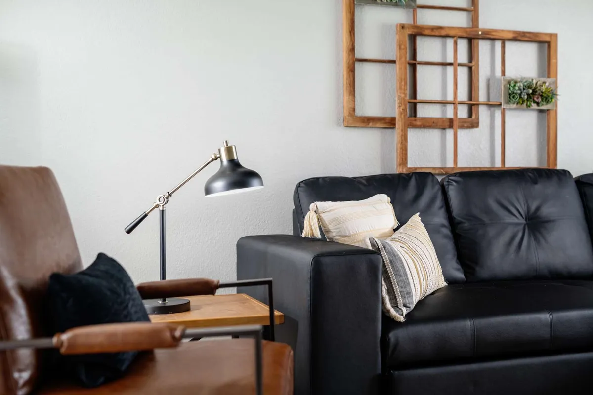 A stylish interior scene featuring a black leather sofa, wooden wall decor, a glowing table lamp, and a wooden side table. The space feels cozy, inviting, and modern, with soft lighting perfect for unwinding after a day of exploring or sharing a romantic evening indoors.