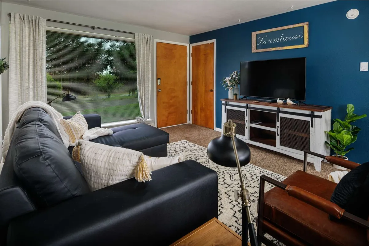 The image showcases the inviting living area of a cozy bungalow, featuring a plush sofa, an armchair, a TV stand, and a large window that bathes the space in natural light. The arrangement offers a perfect spot for relaxing, enjoying movie nights, or curling up with a good book, all while basking in the comfort of a welcoming, tastefully decorated interior.
