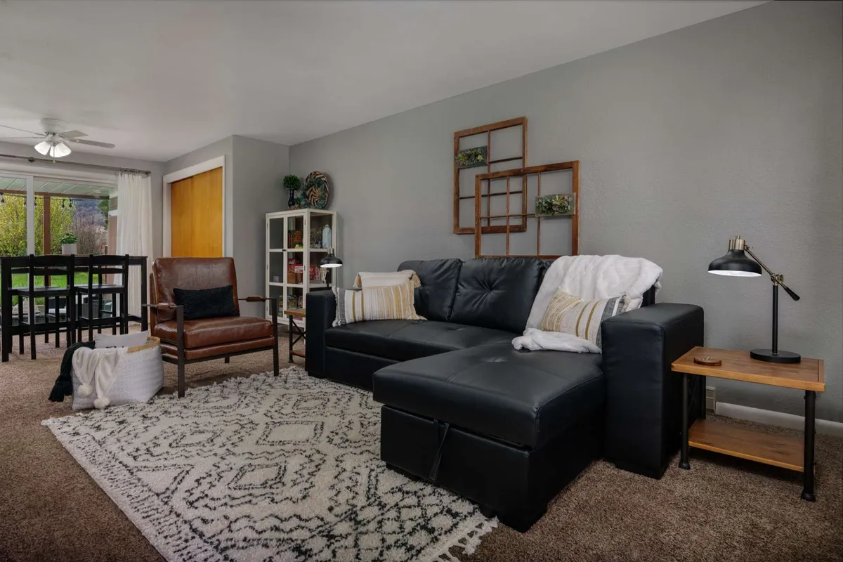 A cozy living room setup with a black sectional sofa, a wooden accent chair, geometric area rug, and decorative wall shelves. The seating arrangement invites relaxation and socializing, echoing the 'heart of the bungalow' ambiance.